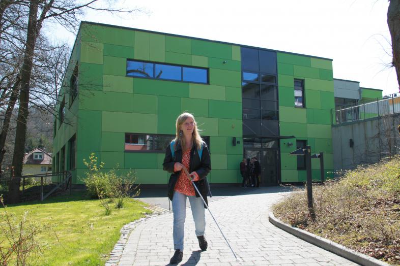 Eine junge Frau mit Blindenlangstock läuft auf einem hell gepflasterten Weg. Im Hintergrund sieht man ein modernes Schulgebäude in verschiedenen Grüntönen. Es ist das Hilde-Klar-Haus auf dem blista-Campus.