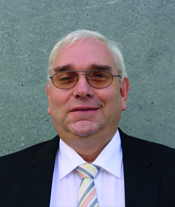 Porträtfoto von Hans-Werner Lange. Vor einem grauen Hintergrund blickt Herr Lange dezent lächelnd direkt in die Kamera. Er trägt ein dunkles Sakko, ein weißes Hemd und eine grau gestreifte Krawatte mit hellgelben und apricotfarbenen Akzenten. Er trägt eine Brille mit getönten Gläsern und kurzes weißes Haar. 